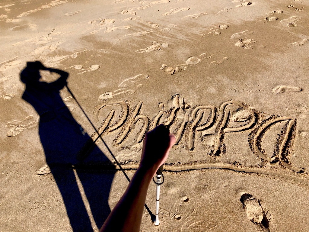 The image above is of a left hand holding a guide cane, looking down onto my name, Philippa, written in the sand. My shadow goes off to the left.