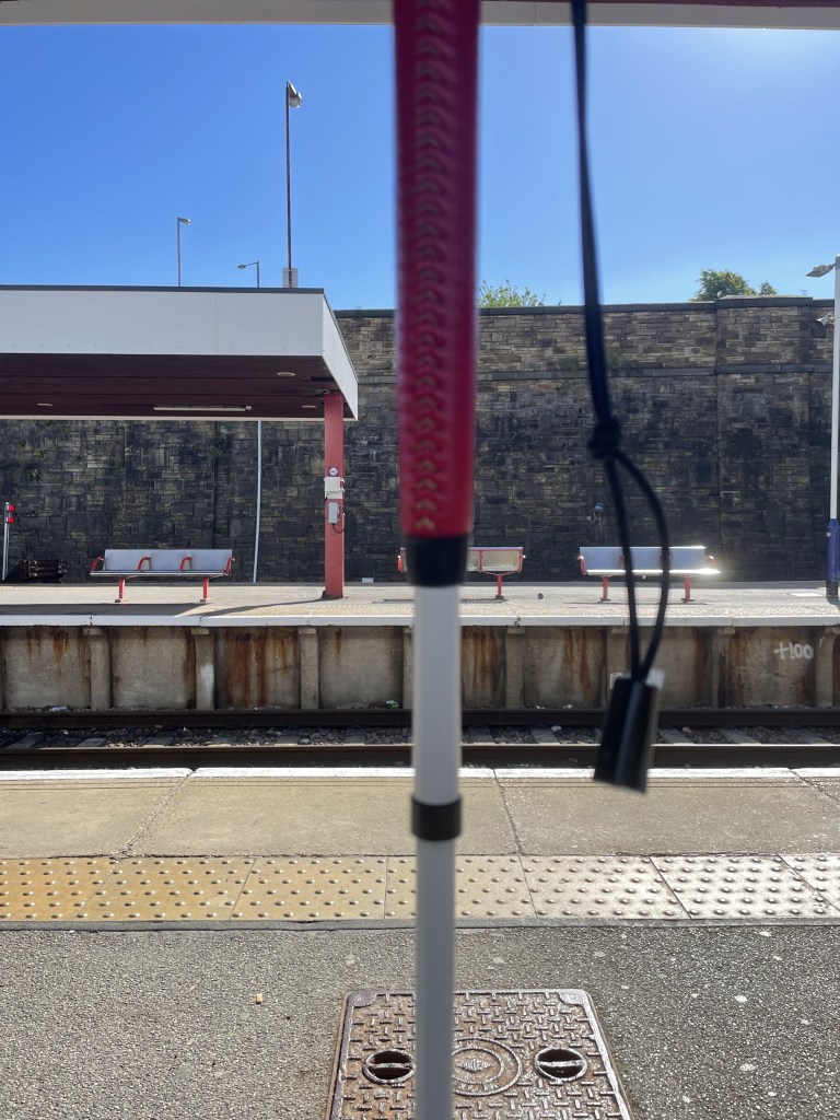 My long cane in the centre, a red handle. In front is a train platform. 