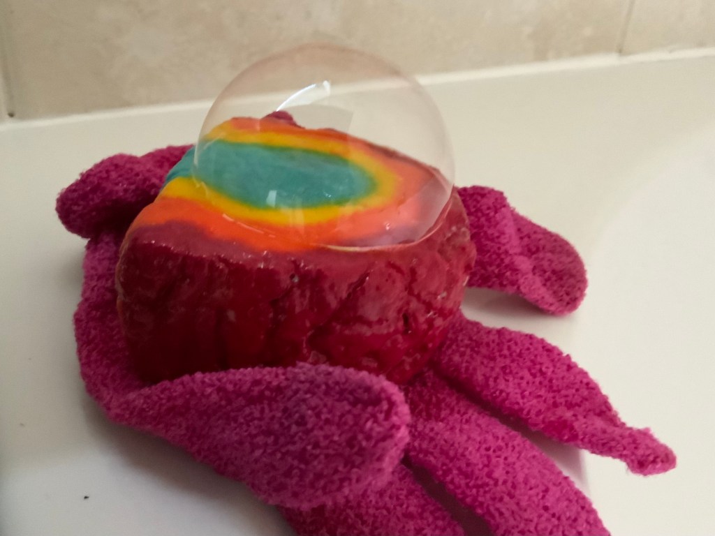 A bubble bar in the colours of the rainbow. Sat on a pink exfoliating glove. There is a bubble on top of the bubble bar. 