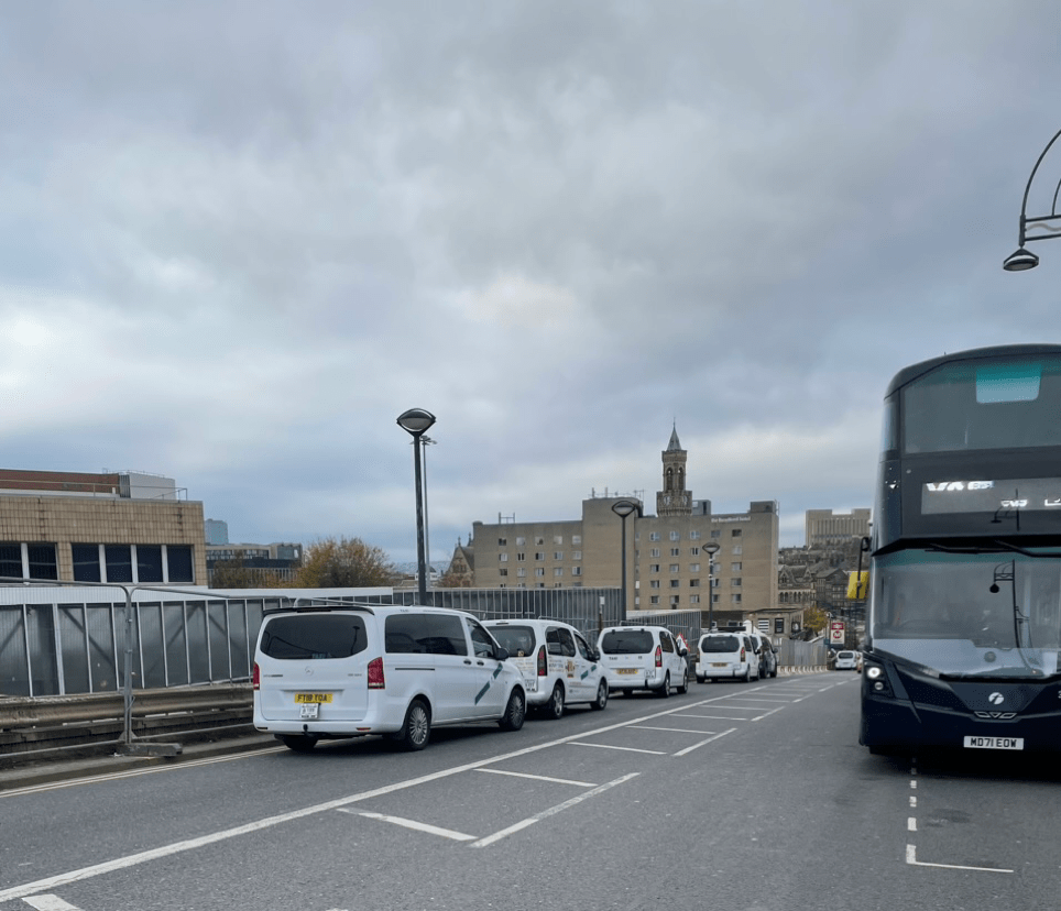Looking down a hill with taxis on one side, with no usable path. A bus is parked on the other side facing up the hill. 