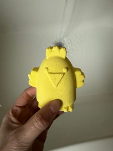A yellow bath bomb shaped like a chicken. Being held over a running bath. 