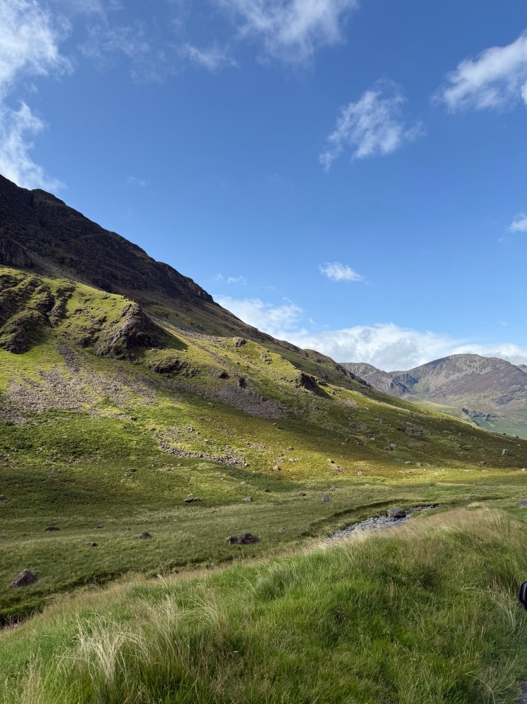 Mountains: a mountain coming down to the left from the right hand side. A small number of mountains in the distance. Th terain is grassy with rocky peaks. The sky isn a light blue with wispy clouds. 